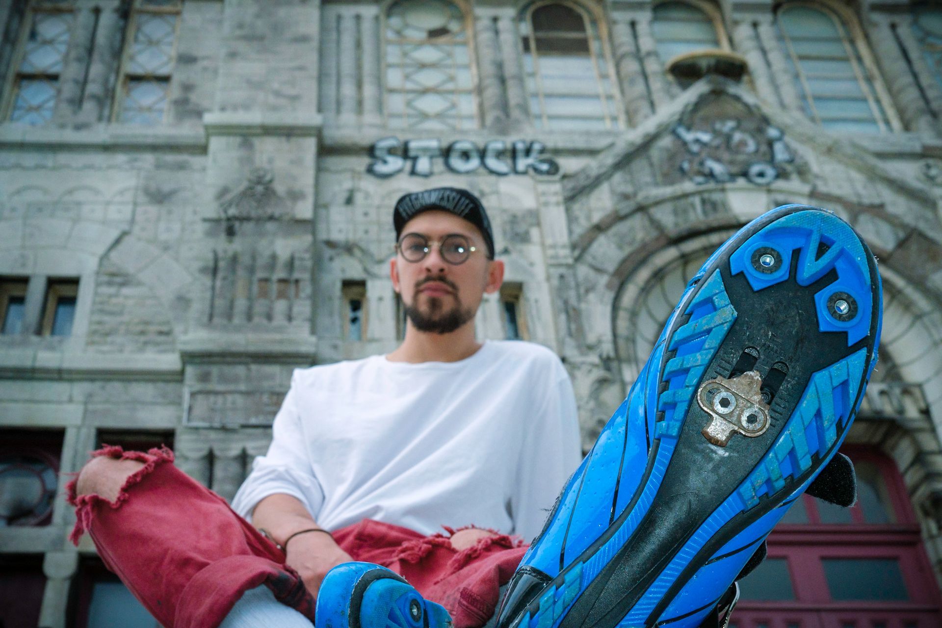Man with glasses and a hat sits, holding a blue skateboard. Building with 