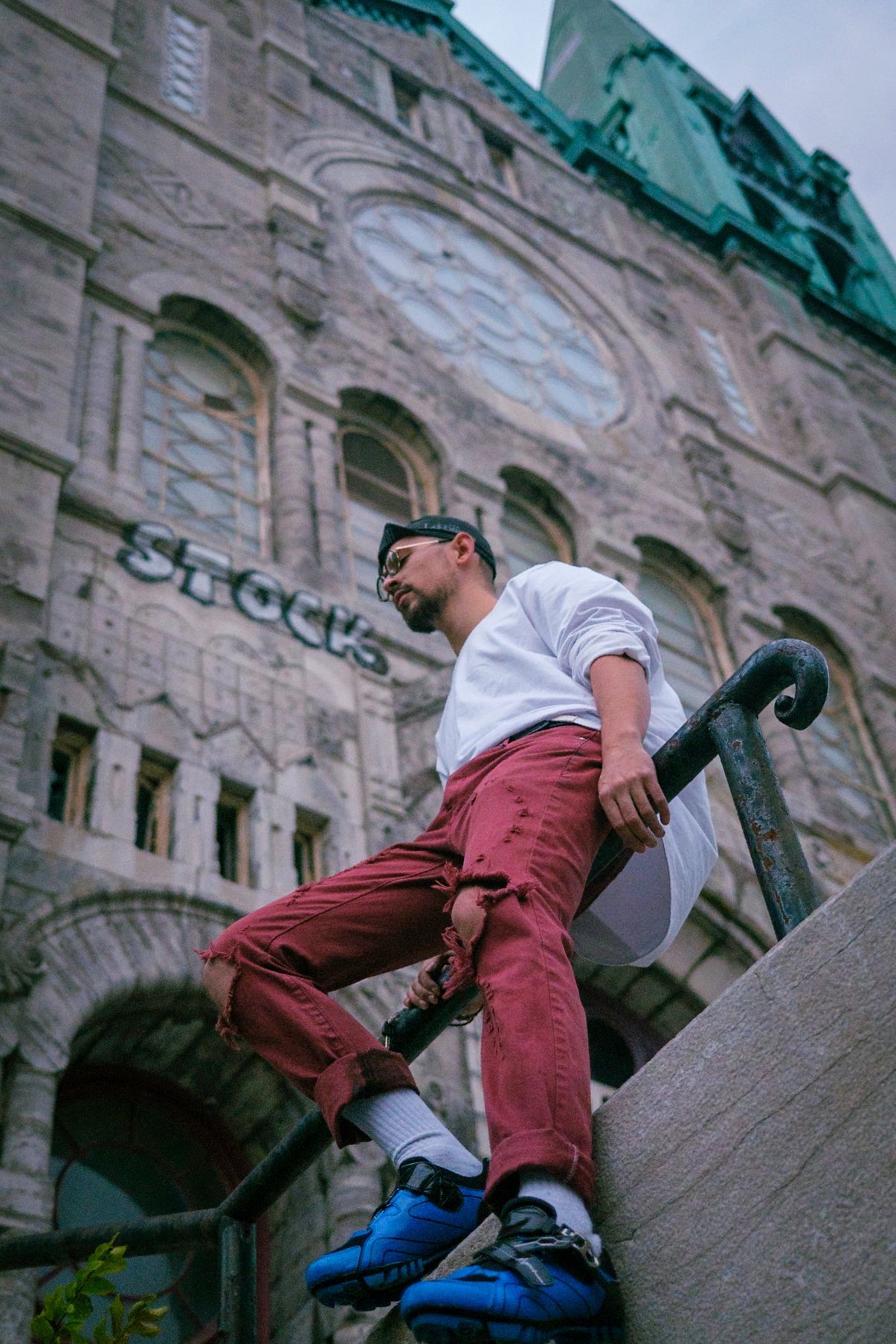 Man in red pants and white shirt leans against a railing in front of a weathered stone building.