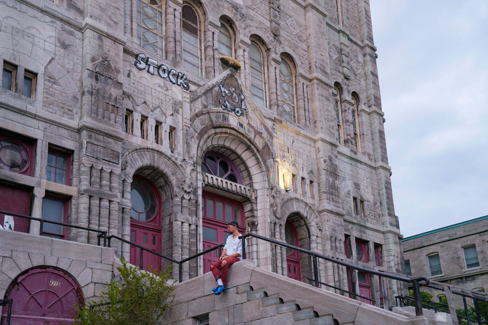 Person sitting on steps of an old, gray stone building with red doors and arched entrance.