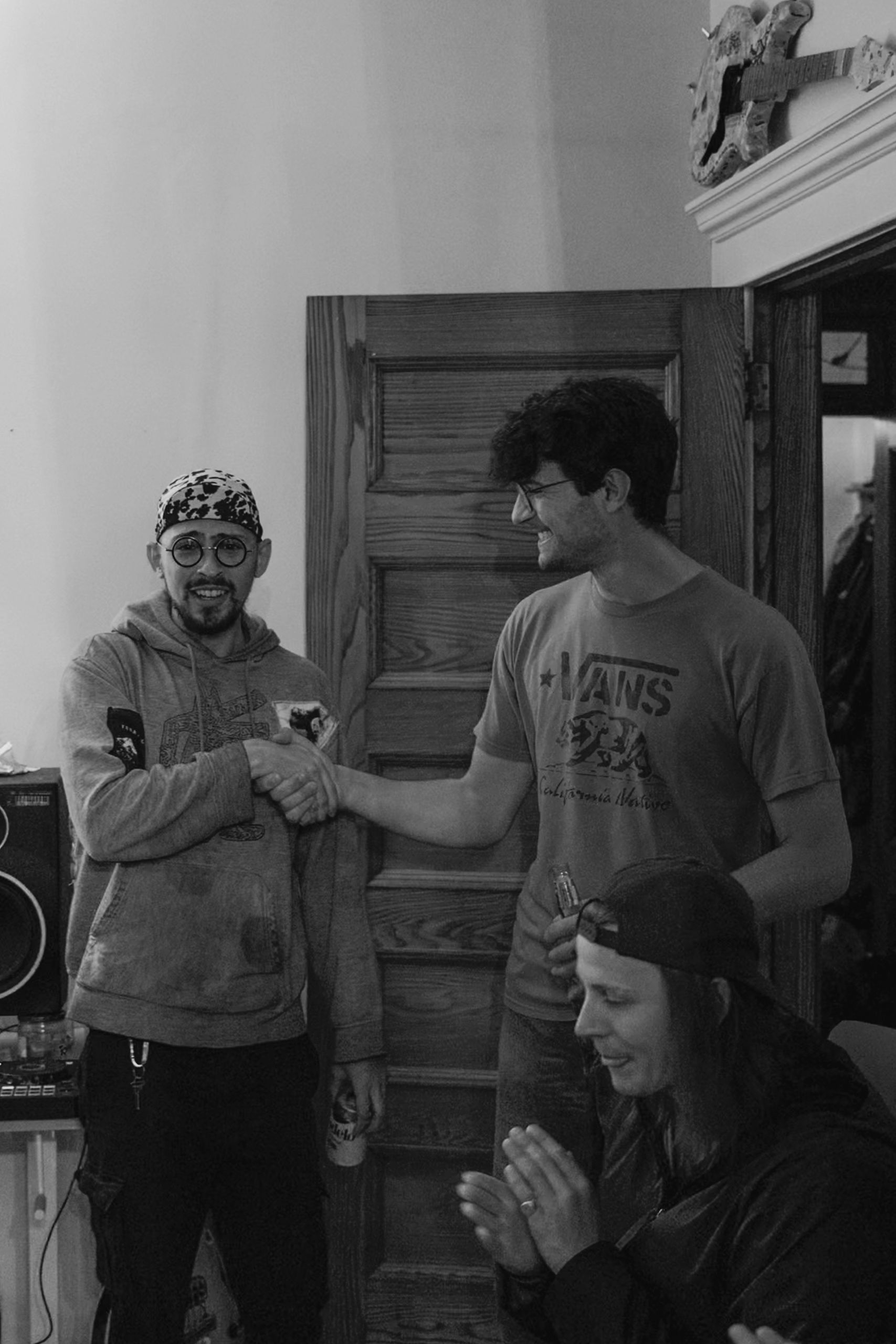 Three people in a room; two shaking hands, one applauding. Wooden door, speaker, and decorative molding visible. Black and white.