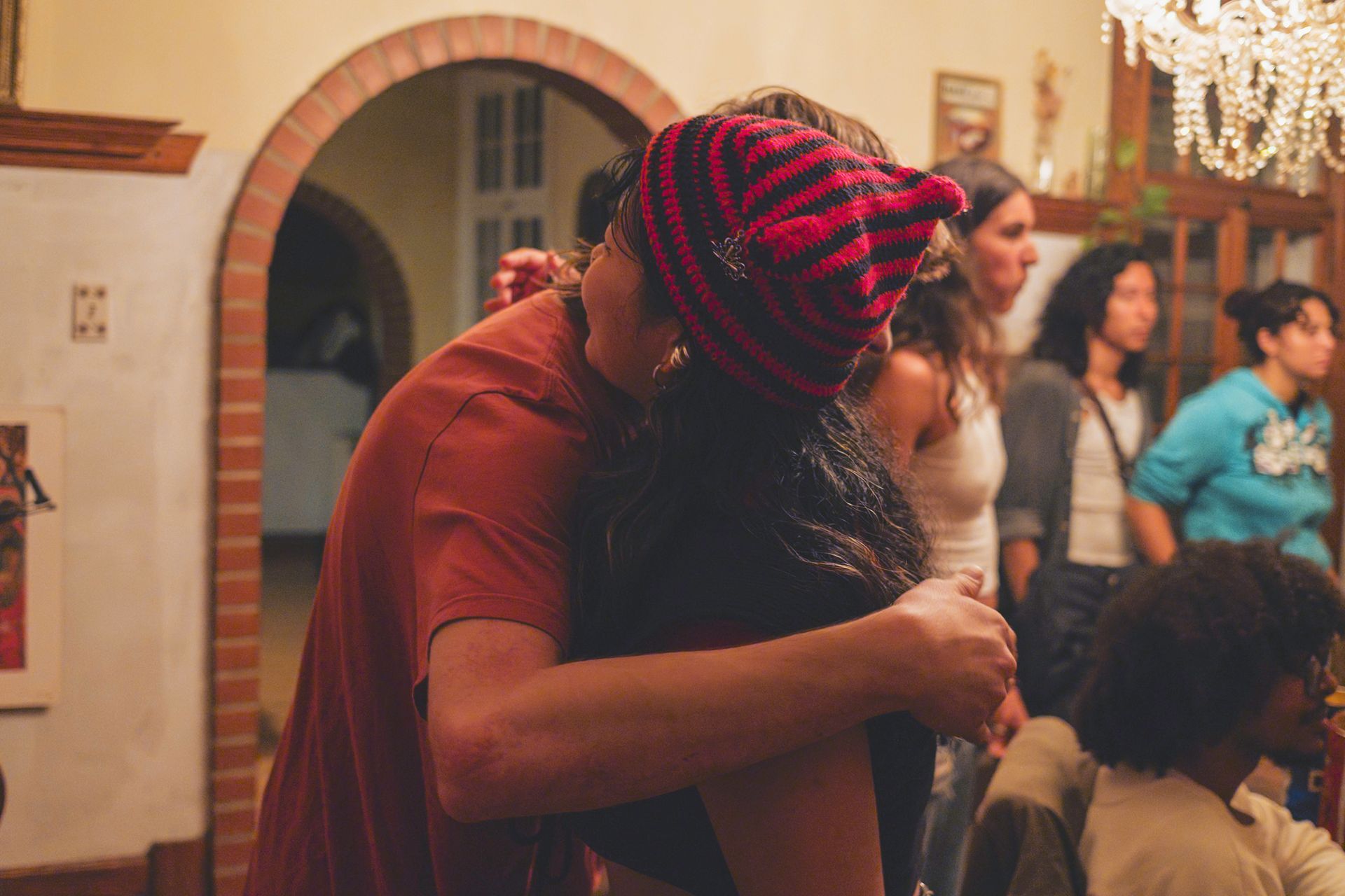 Couple embracing in a room; a red and black beanie, several people watching in background.