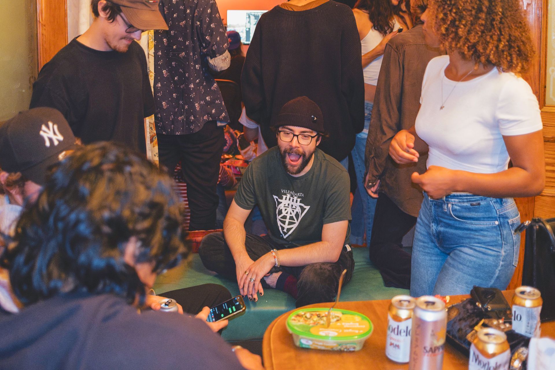 People gathering indoors, some seated on the floor. A man with open mouth sits cross-legged. Food and drinks visible.