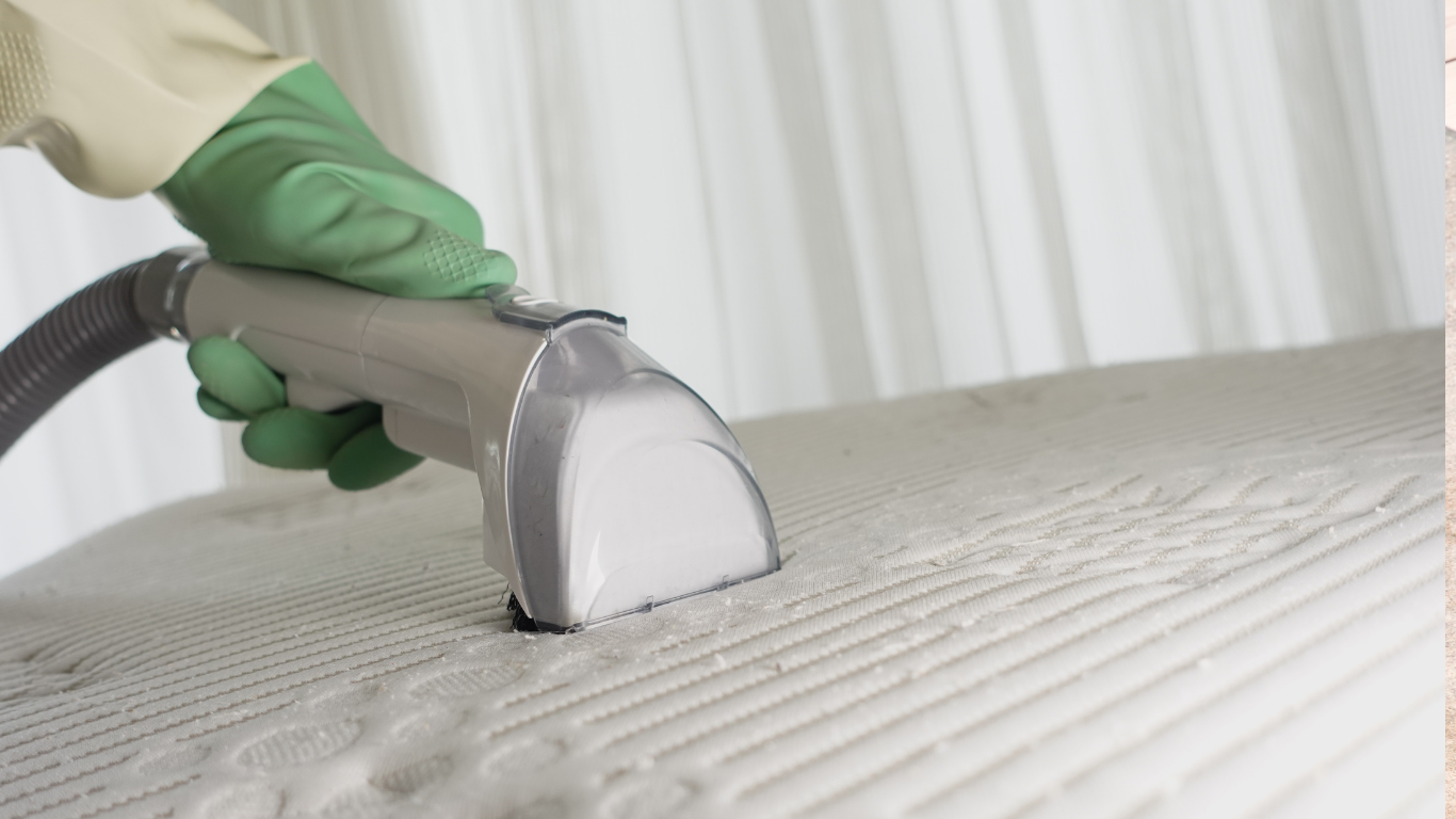 Person in green gloves cleaning a white mattress with a handheld vacuum.