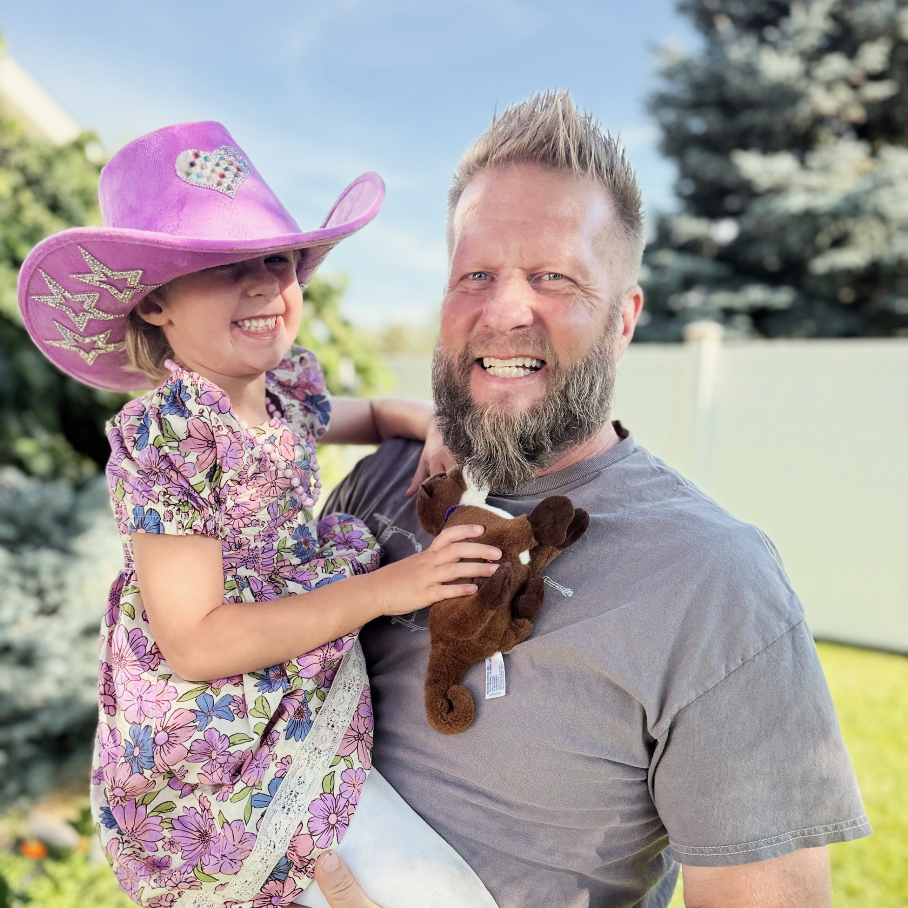 Man holding a smiling girl in a purple cowboy hat. They’re outdoors on a sunny day, both looking at the camera.