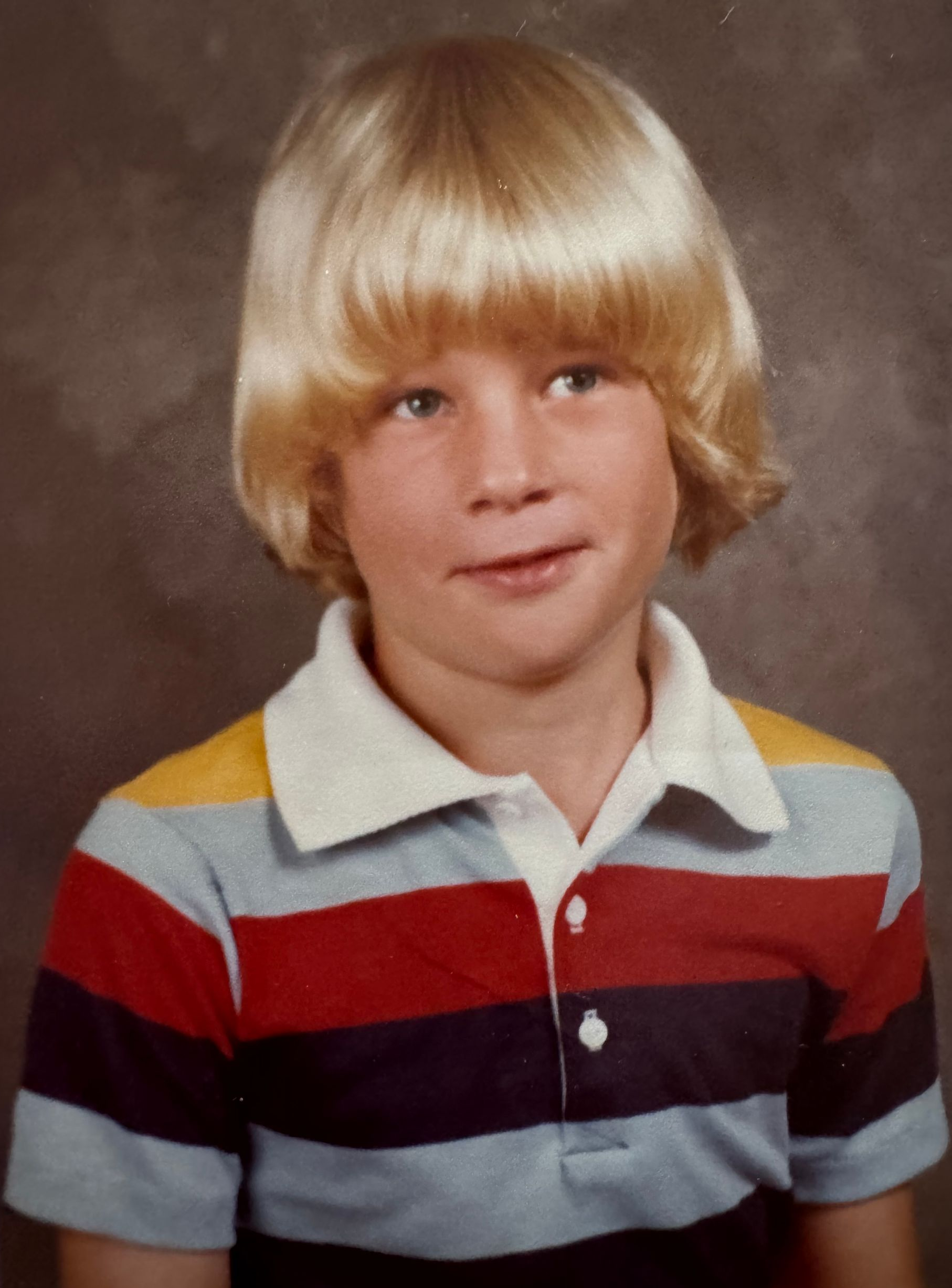 Blond-haired boy in striped shirt, smiling, school portrait.