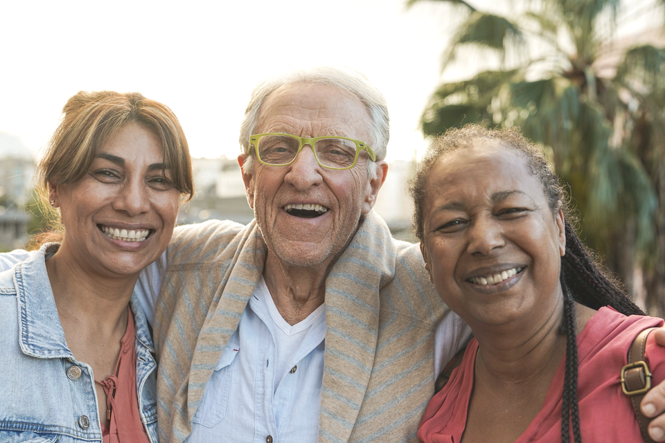 Three smiling people: an older man with glasses between two women outdoors.
