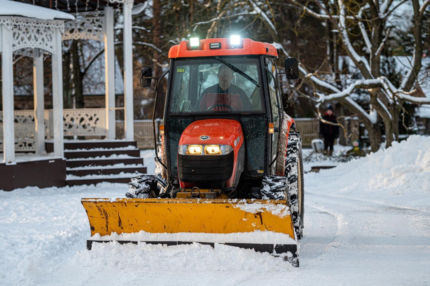 Orange tractor plowing snow, outdoors. Bright lights, yellow blade, trees and building in the background.