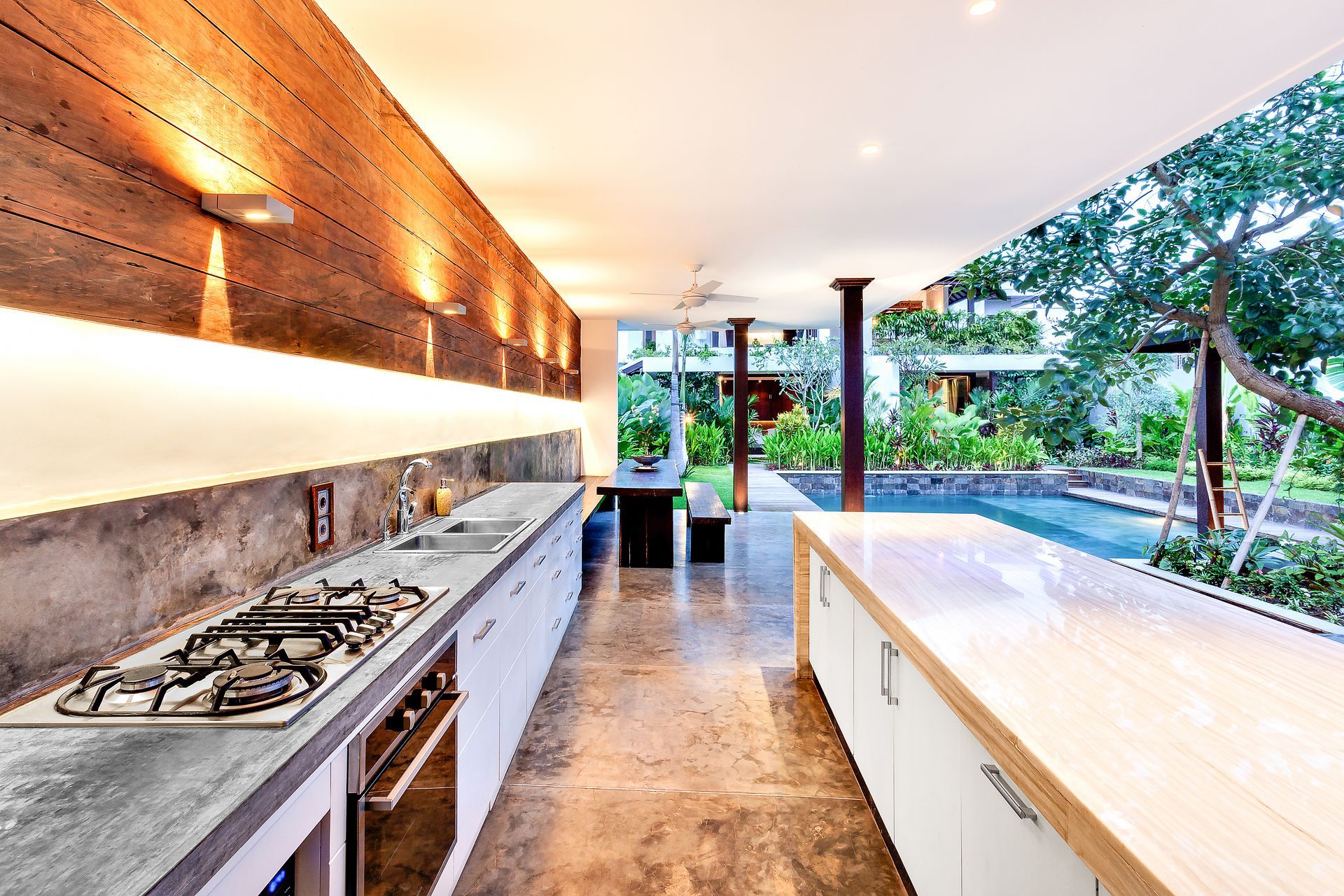 Modern outdoor kitchen with gas stove, long counter, and open view to a pool and garden.