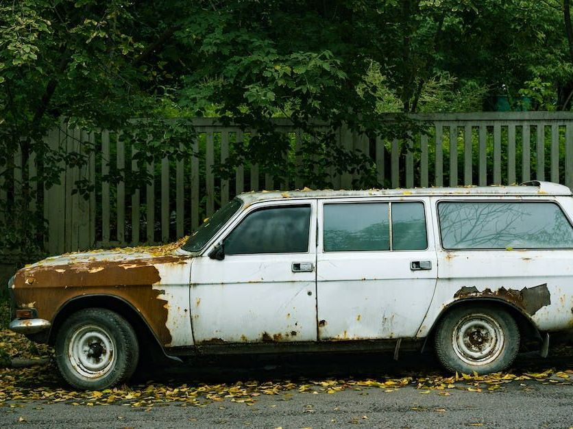 A rusty white car is parked on the side of the road.