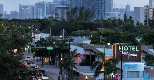 Street view with a Hotel New Yorker sign, cars, trees, and tall buildings in the background at dusk.