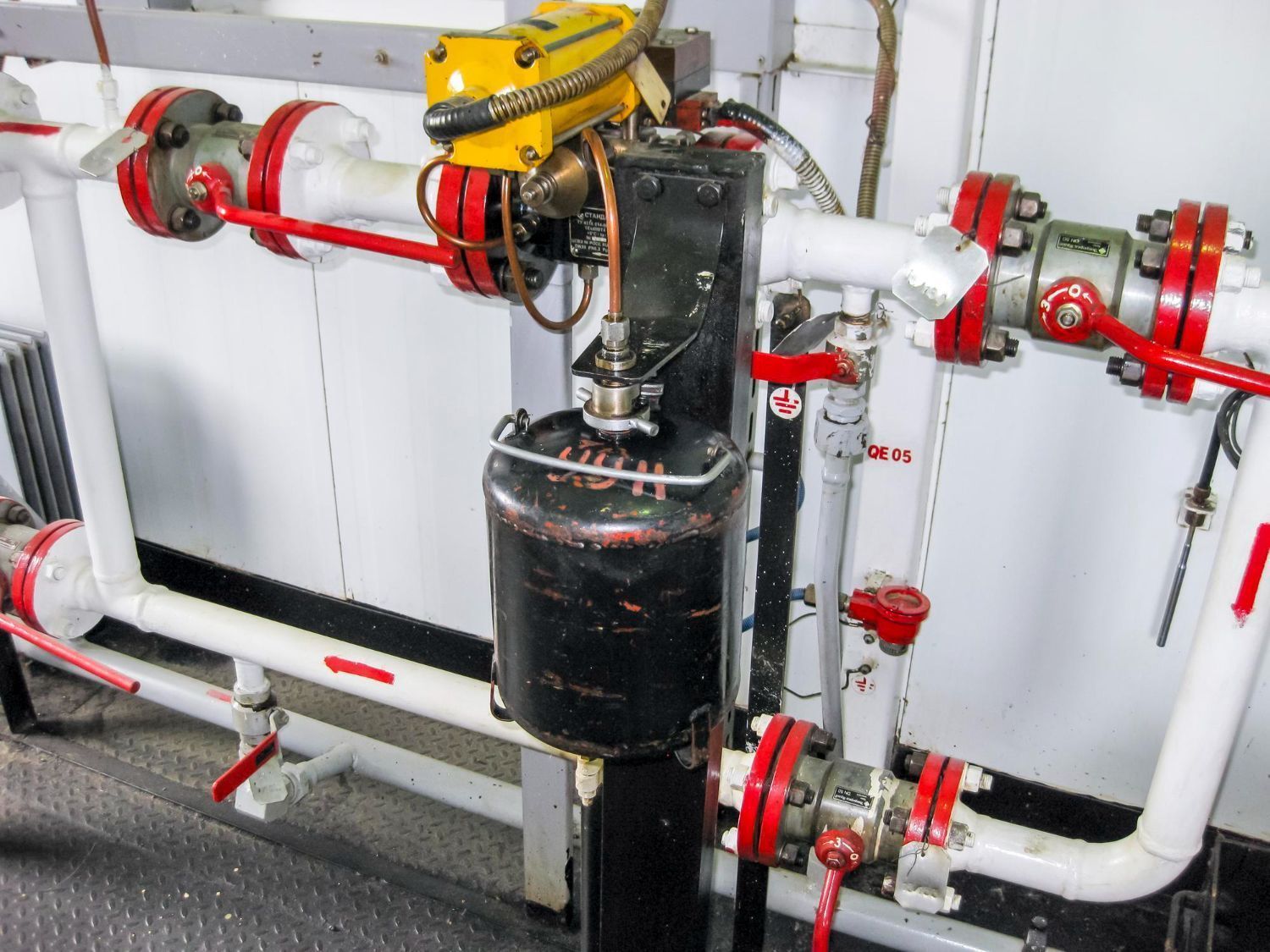 Industrial piping with red valves and a black tank-mounted valve assembly in a mechanical room