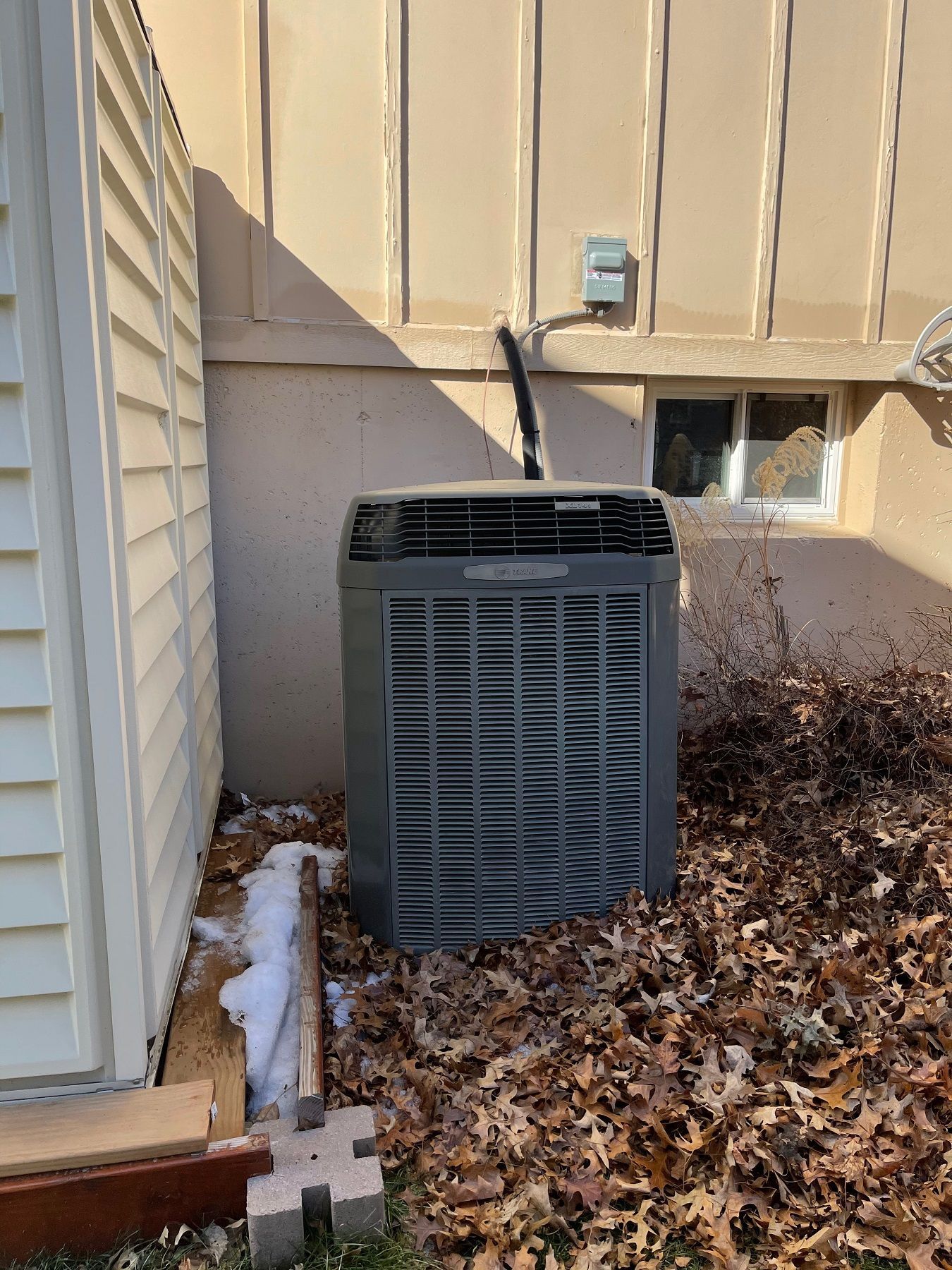 Outdoor air conditioner unit beside a beige house wall with patchy snow and dead leaves nearby