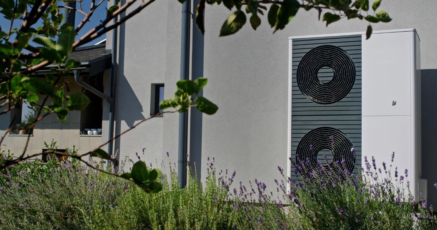 Outdoor heat pump unit beside a building, partly hidden by tall grass and leafy branches.