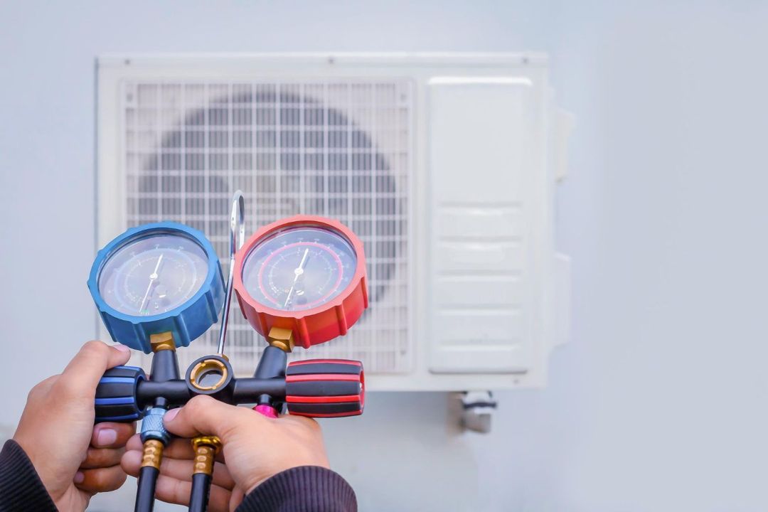 Hands holding blue and red manifold gauges connected to an outdoor air conditioner unit