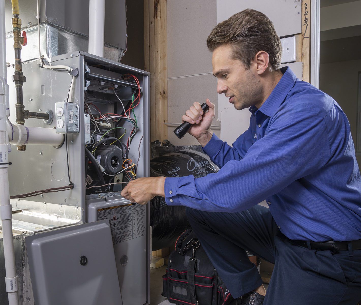 Technician in blue shirt inspecting open furnace wiring with a flashlight in a utility room