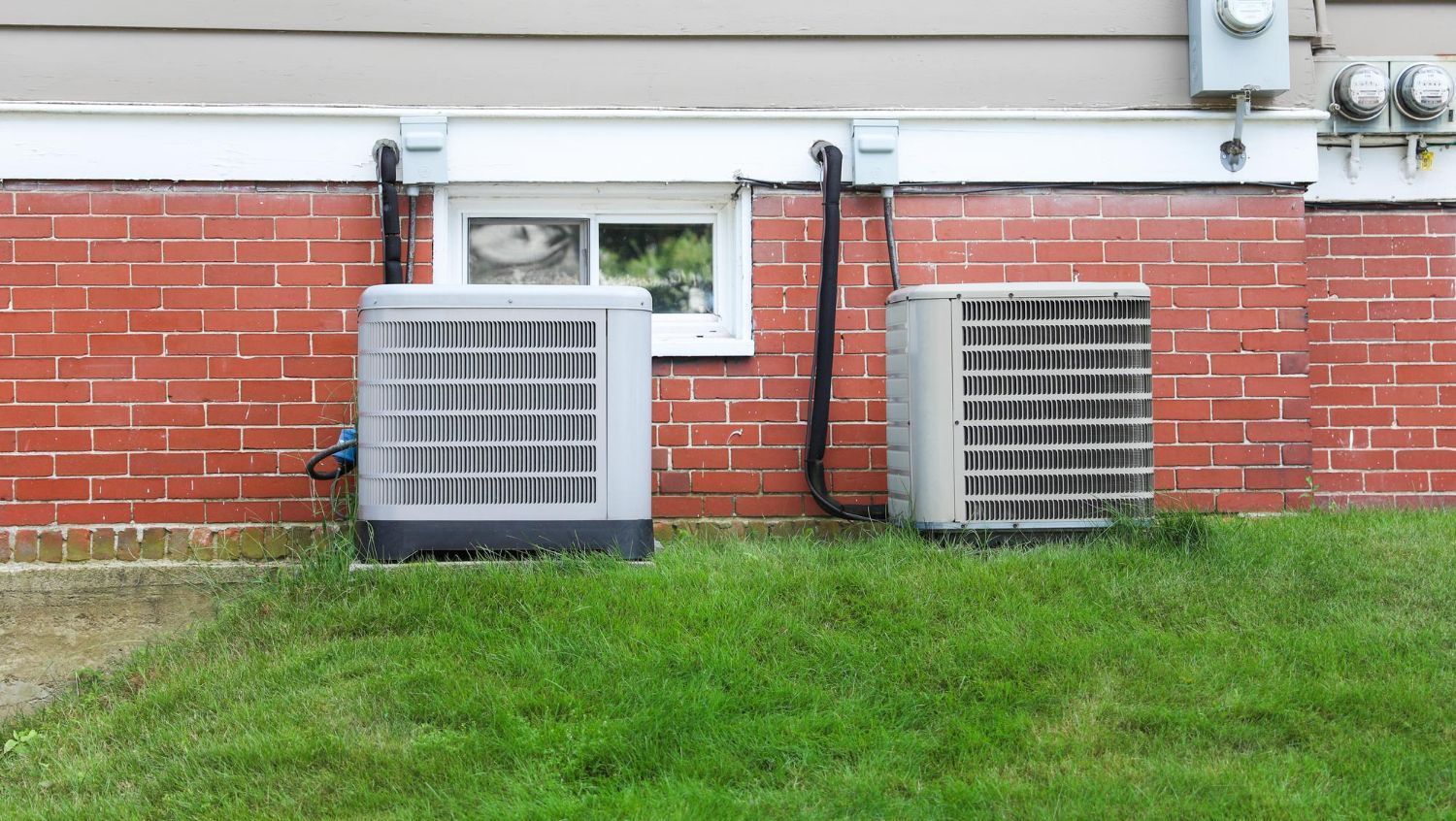 Two large gray HVAC units on grass beside a red brick building wall with a small window.