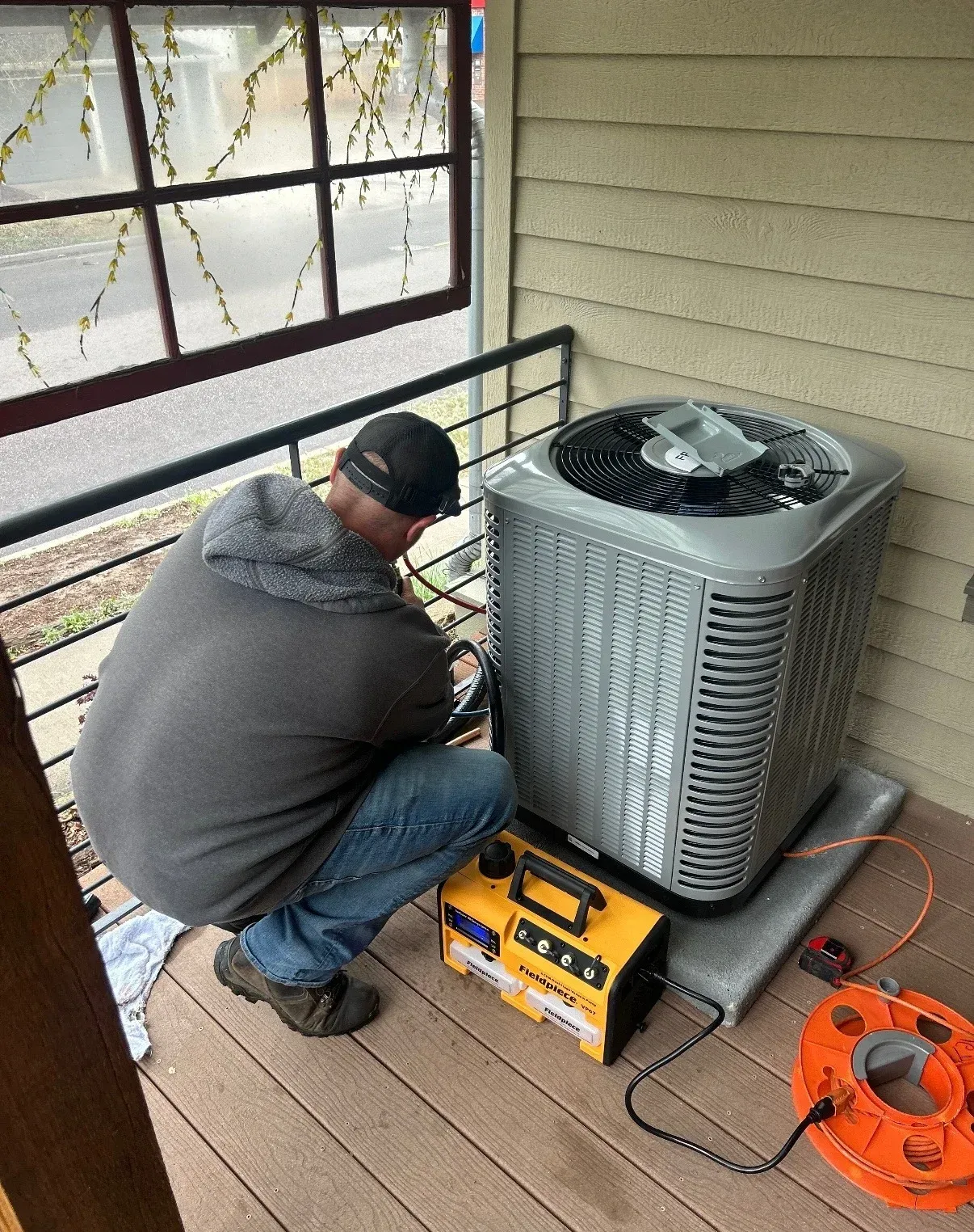 Technician kneeling beside a rooftop air conditioner, using a yellow service tool and orange hose reel.