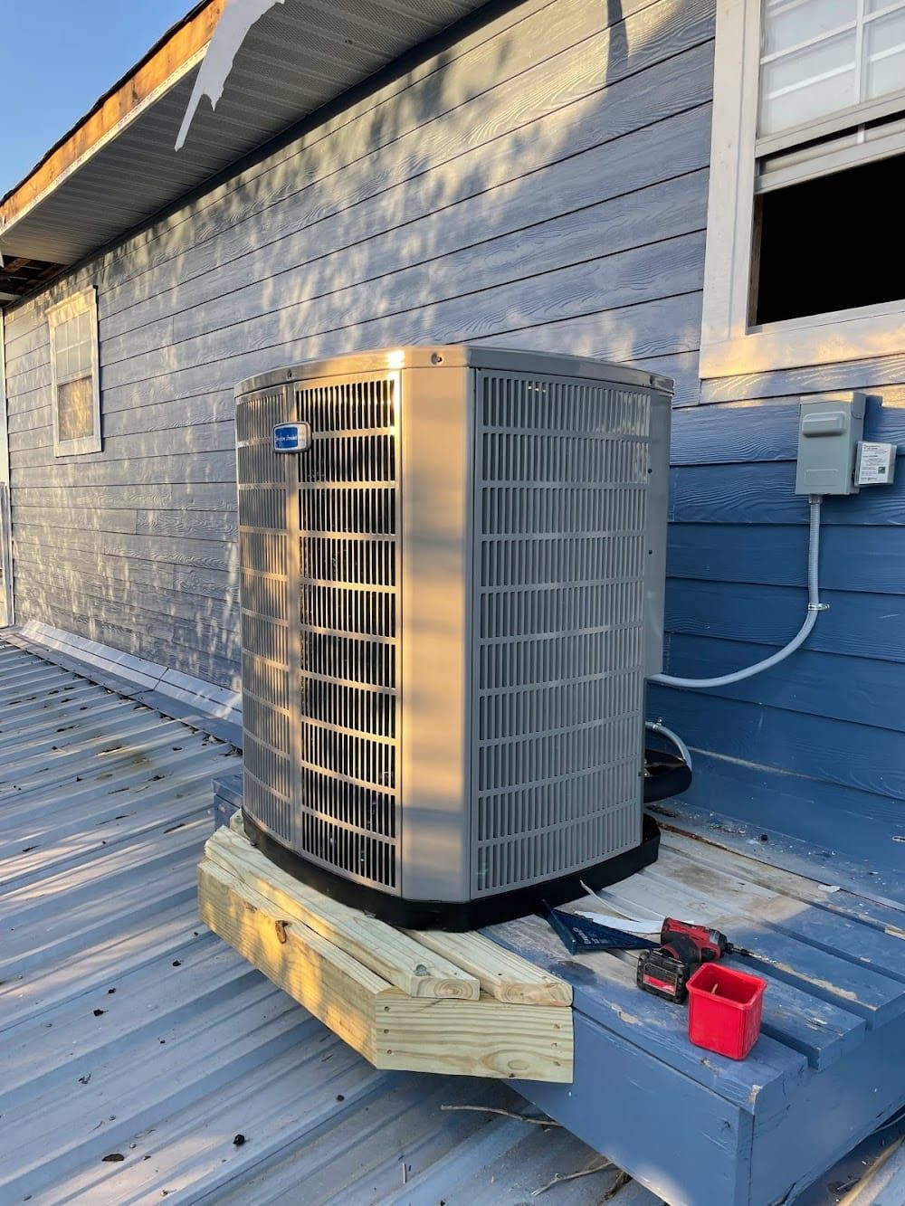 Outdoor HVAC unit on a rooftop beside a blue wall and window, with red tool case nearby