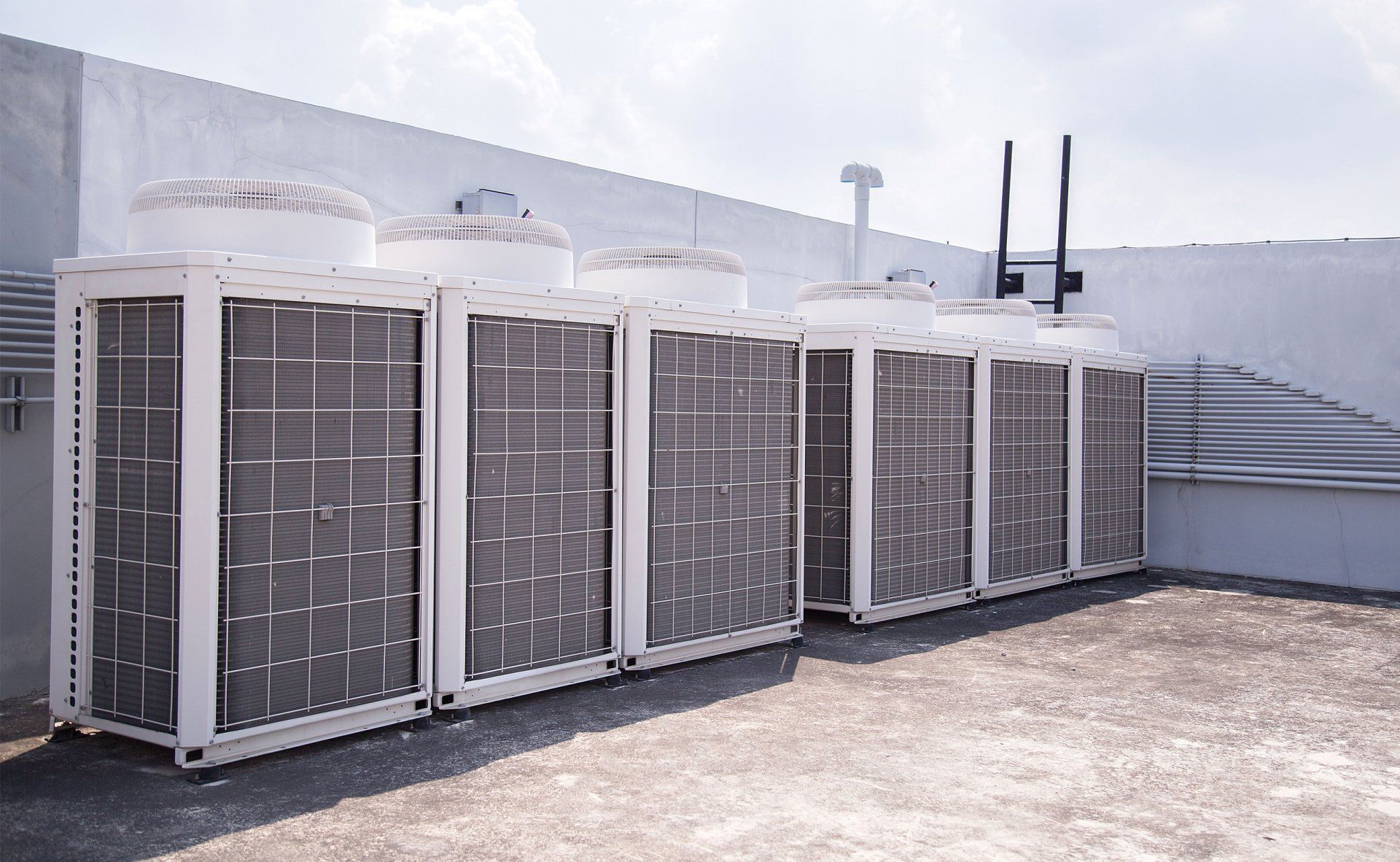 Row of outdoor HVAC units with metal grilles beside a white building on a rooftop or industrial site