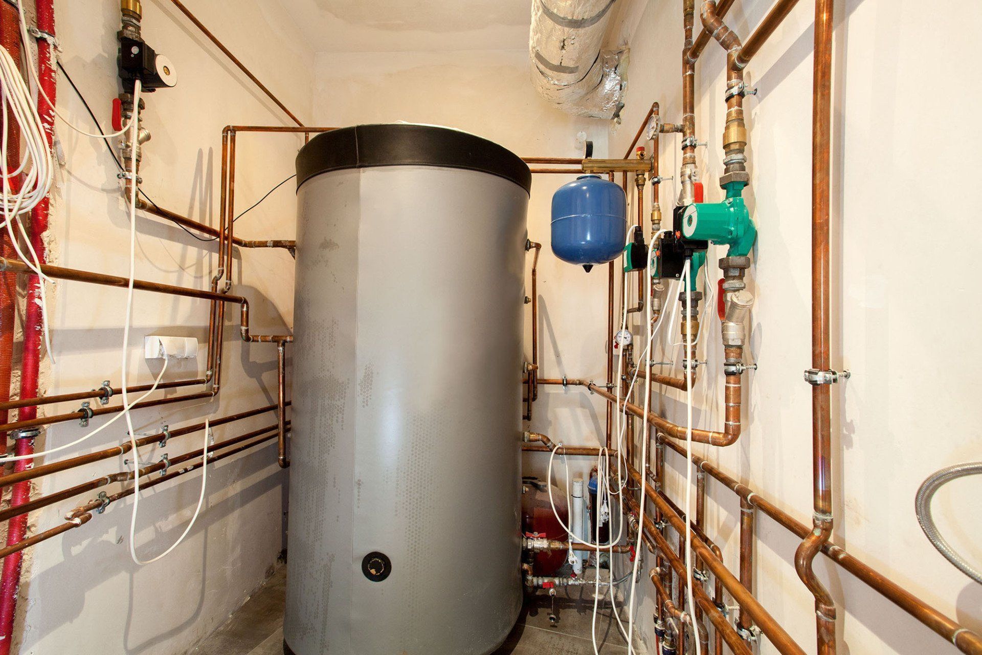 Boiler room with a large gray water heater, exposed copper pipes, and wall-mounted valves and tanks