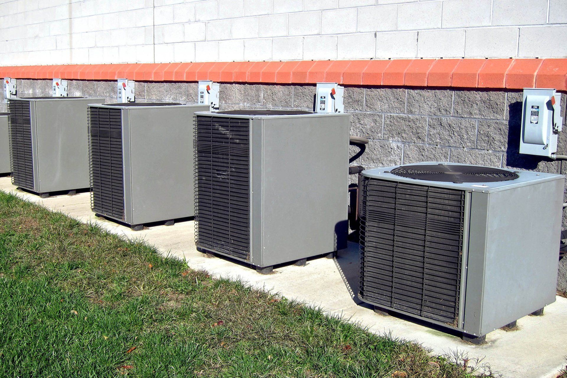 Four outdoor HVAC units beside a building wall on a concrete pad рядом with grass