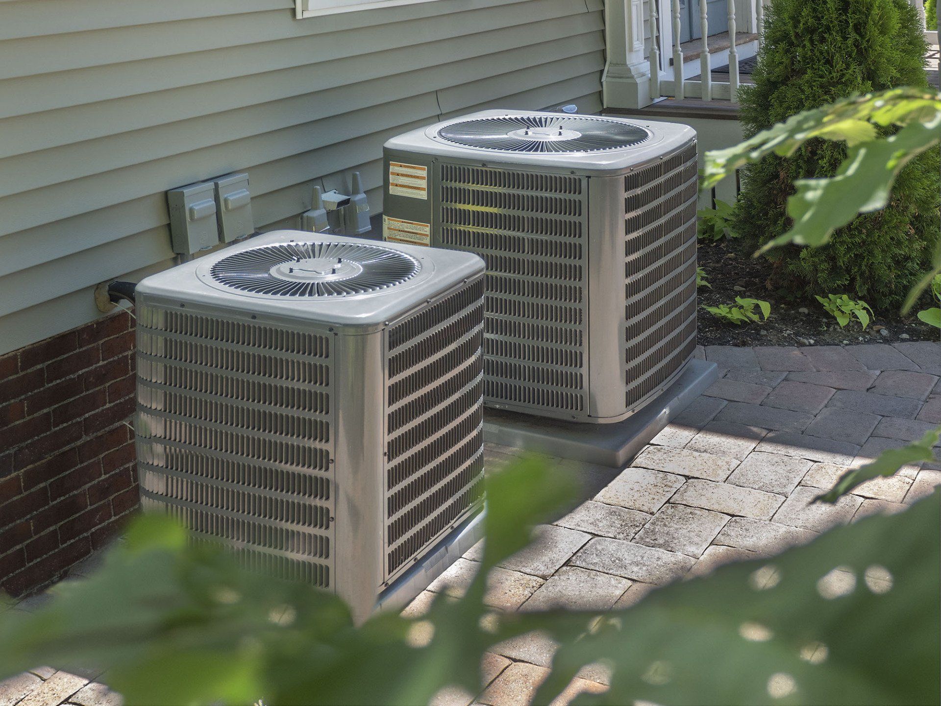 Two outdoor air conditioning units beside a house wall, partly framed by greenery