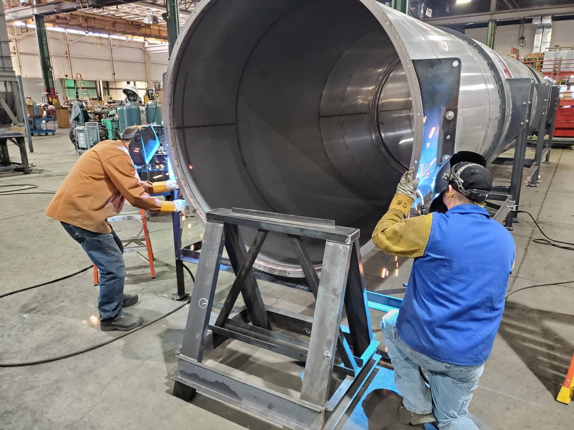 Workers assembling a large metal industrial cylinder in a warehouse workshop