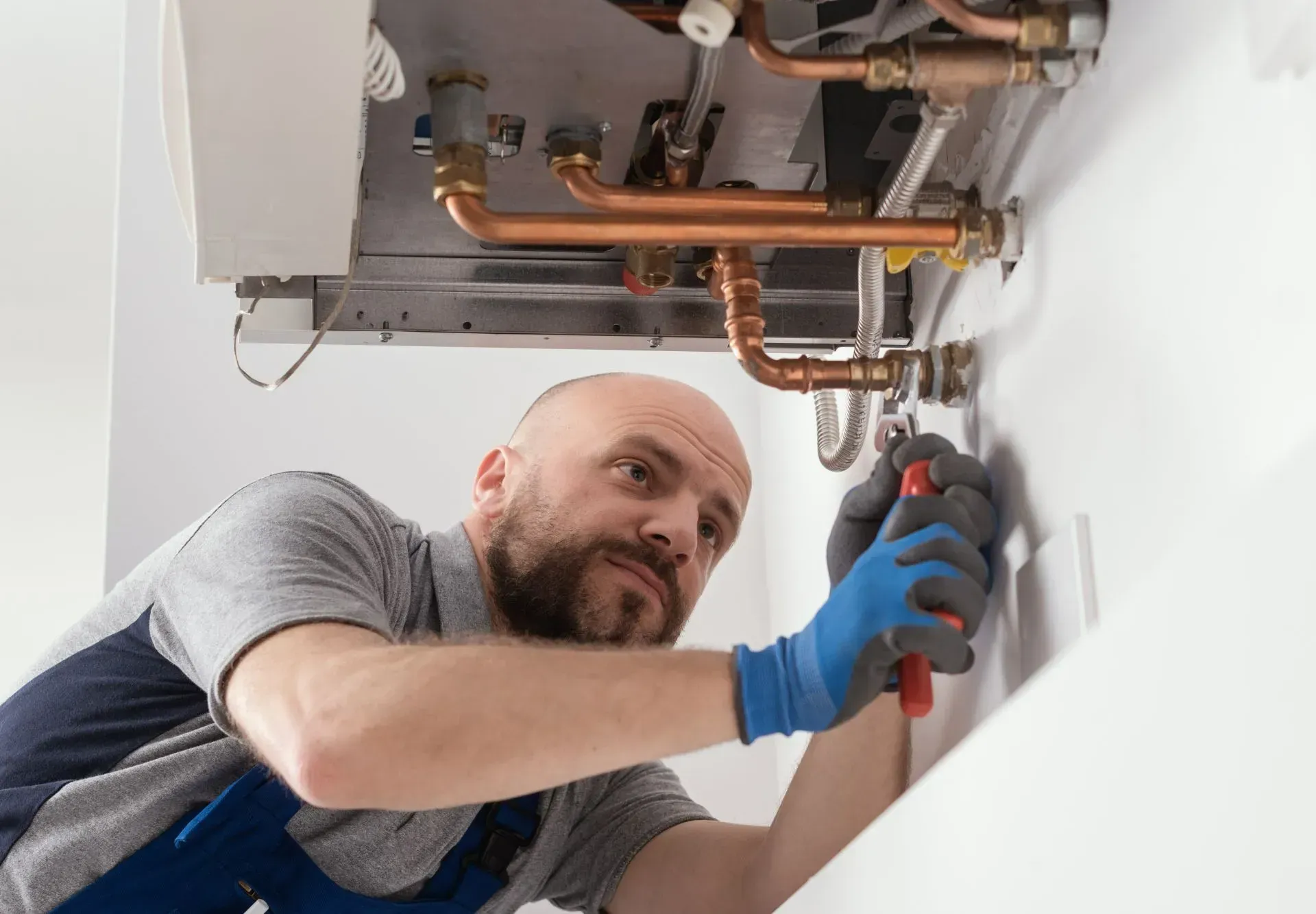 Plumber using a wrench to repair exposed pipes under a sink, wearing blue gloves.