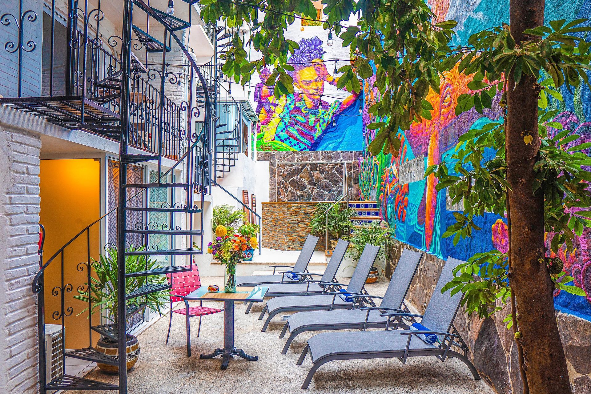 A patio with chairs , tables and a spiral staircase with a colorful mural on the wall.