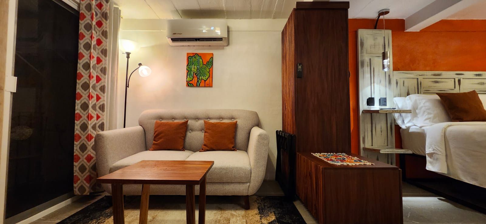 A cozy studio apartment with a beige sofa, orange accent wall, and wooden furniture.