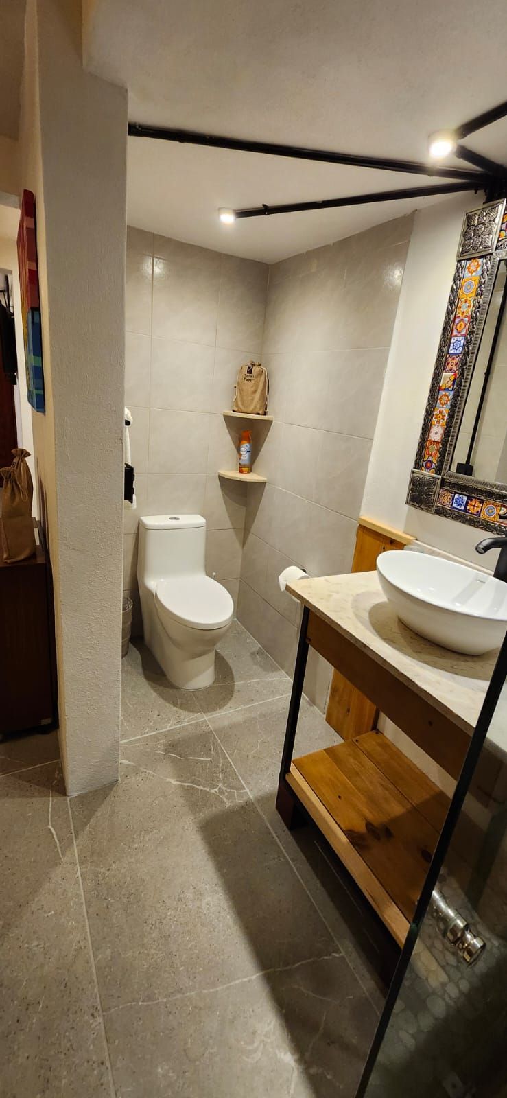 A bathroom with a toilet, a sink, and a wooden shelf. Gray tiled walls and floor.