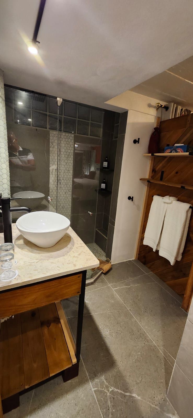 Bathroom with a white sink on a wooden vanity, glass shower, wooden shelf with towels, and gray tile floor.