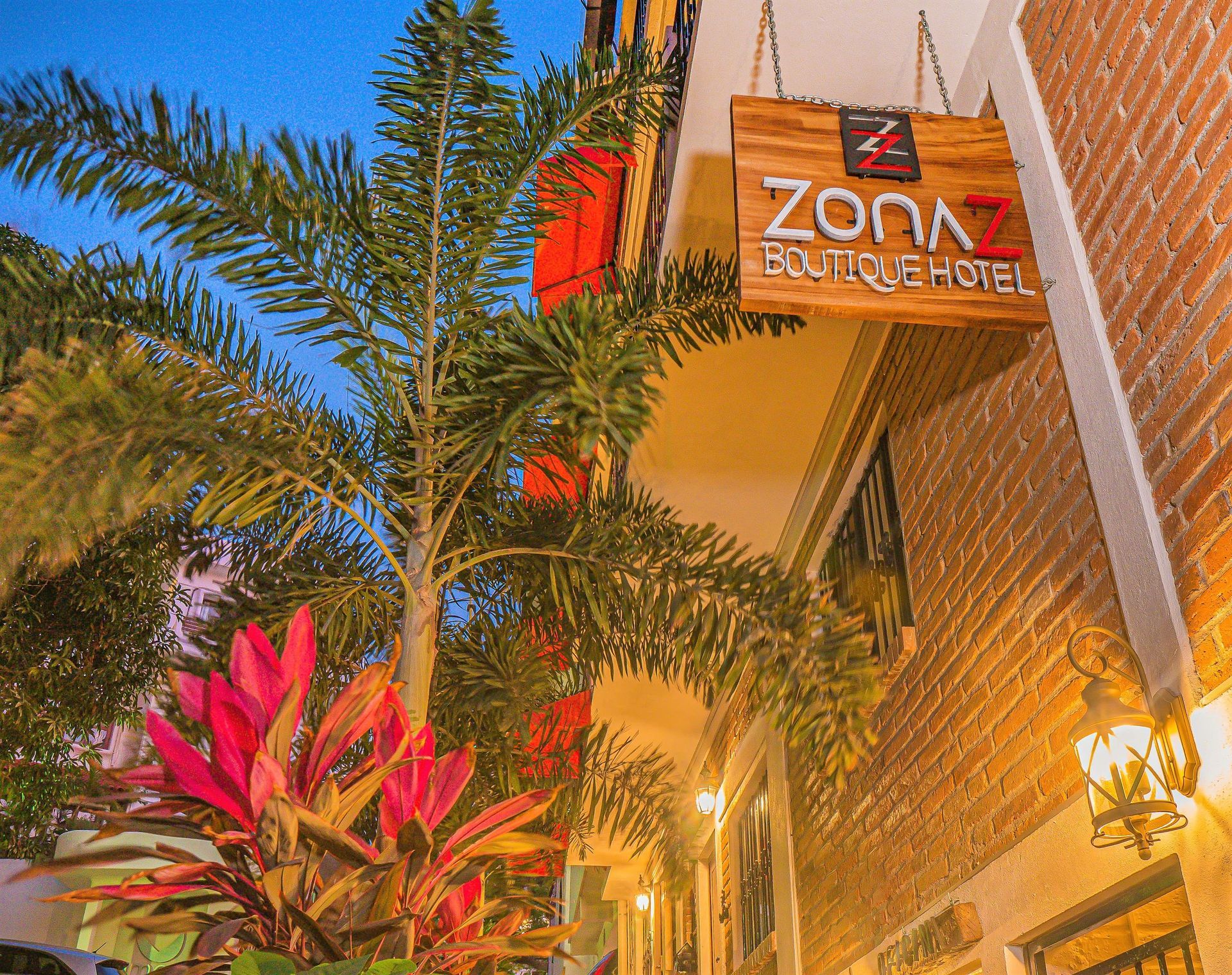 Wooden sign for Zona 7 Boutique Hotel, palm tree, brick wall, and hanging lantern.