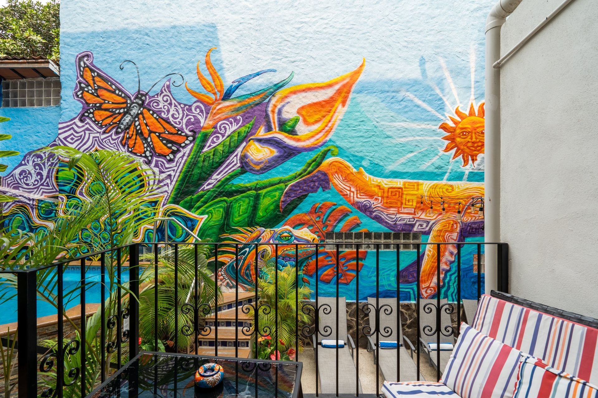 Mural of a butterfly, flowers, and sun on a building, viewed from a patio with black railing, table, and striped pillows.