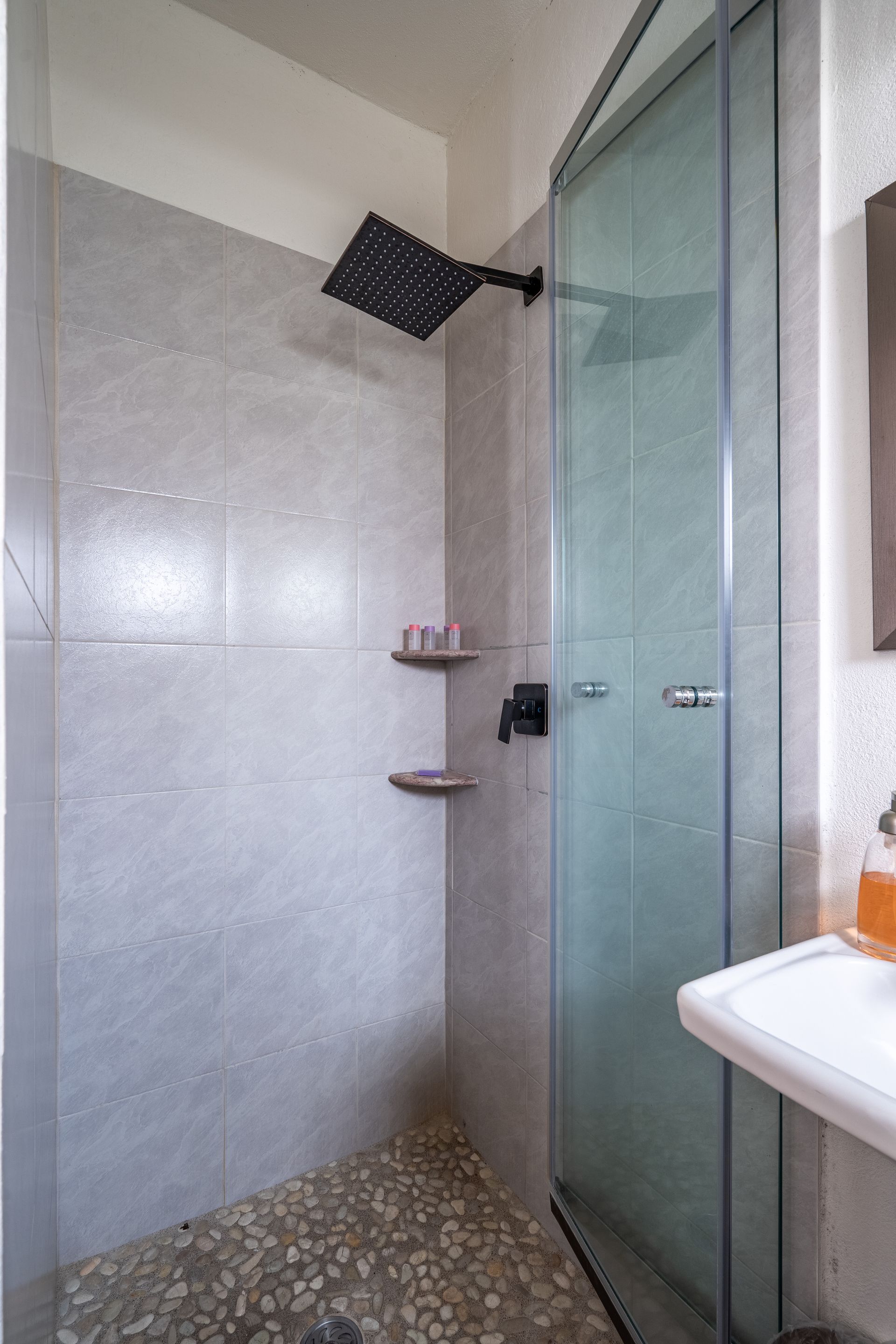 Shower with pebbled floor, gray tile walls, glass door, and dark fixtures.