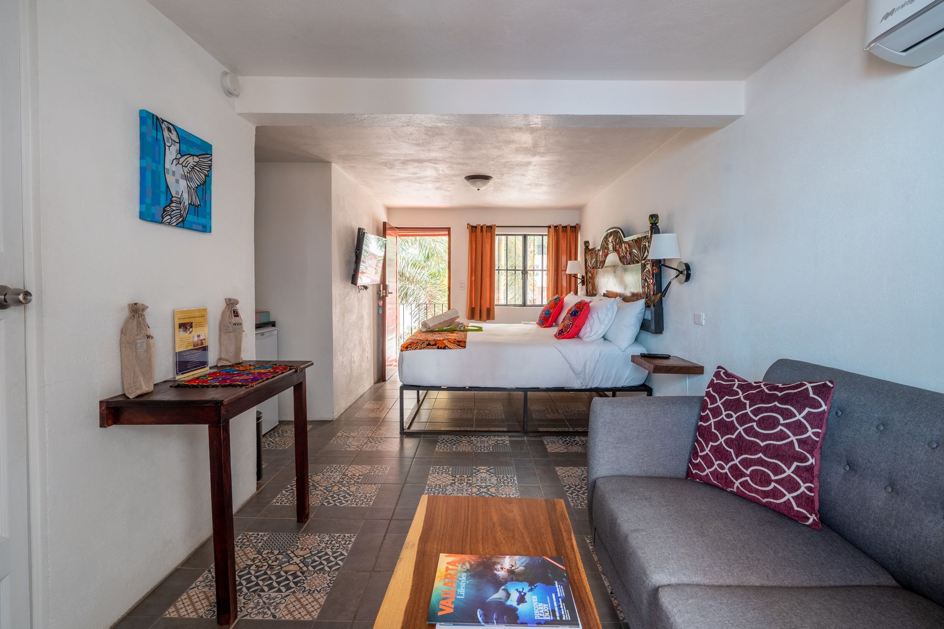 Cozy studio apartment with gray sofa, bed, and wooden desk. Bright natural light through the open doorway.