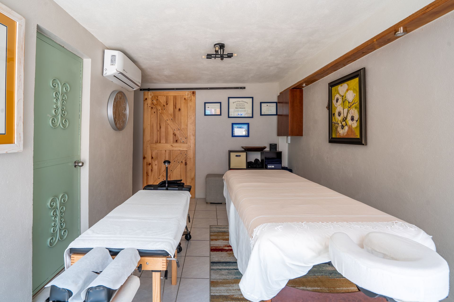 Two massage tables in a therapy room. Wooden door, light green door, awards on wall, and flowers.