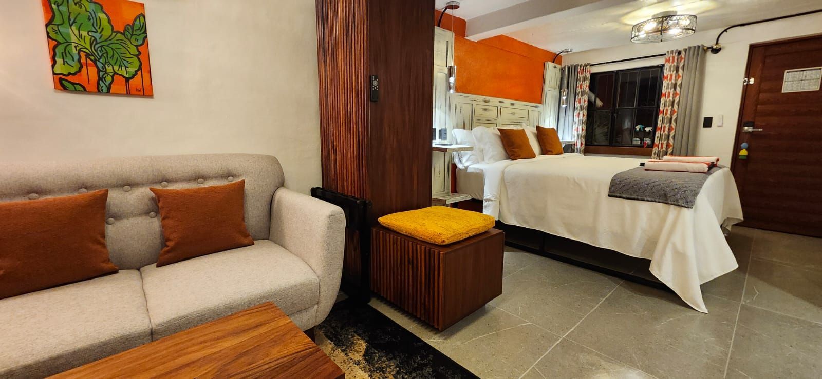 A small, well-lit hotel room with a bed, couch, and wooden storage. Earth-tone colors throughout.