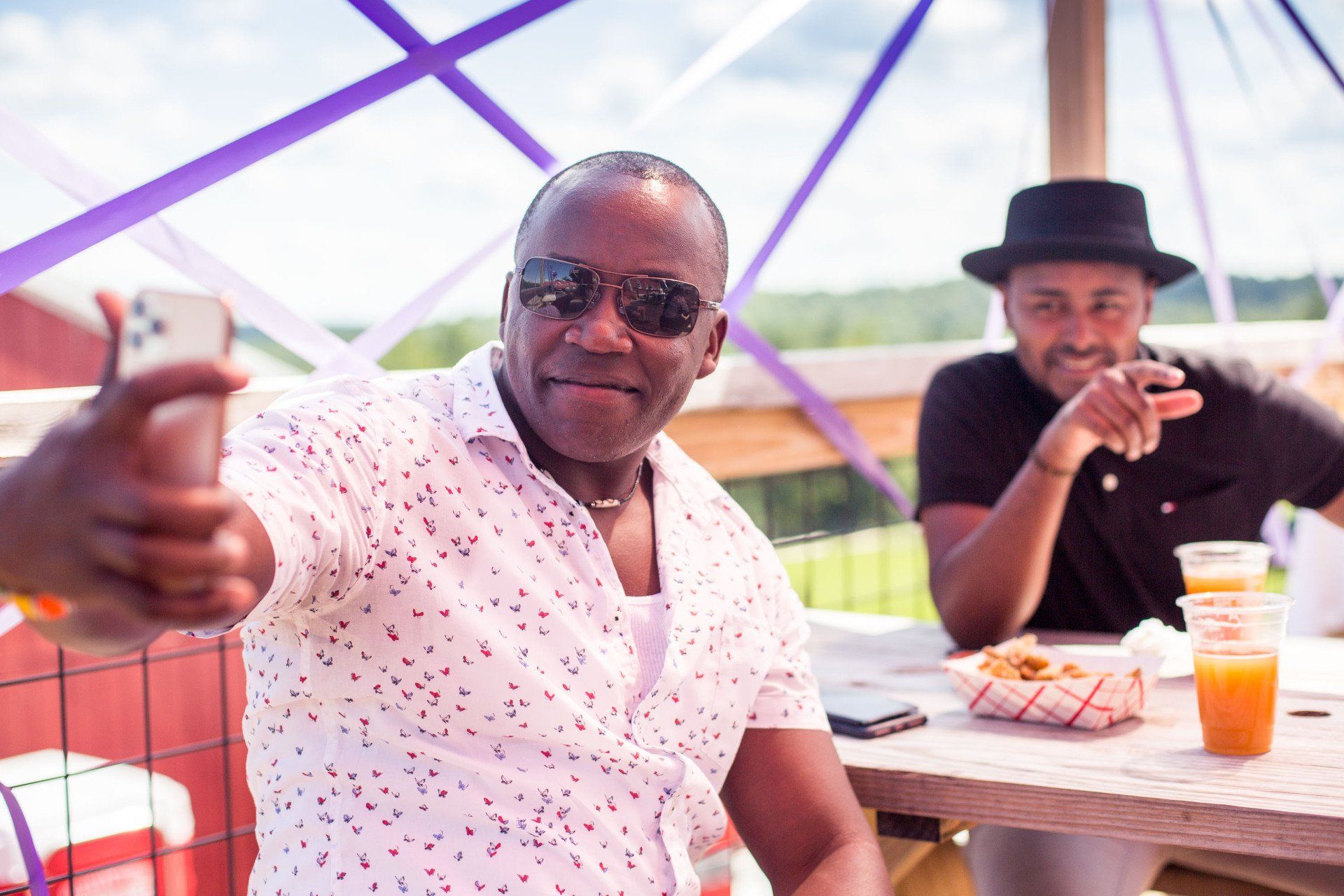 Two men are sitting at a picnic table and one of them is giving a thumbs up.