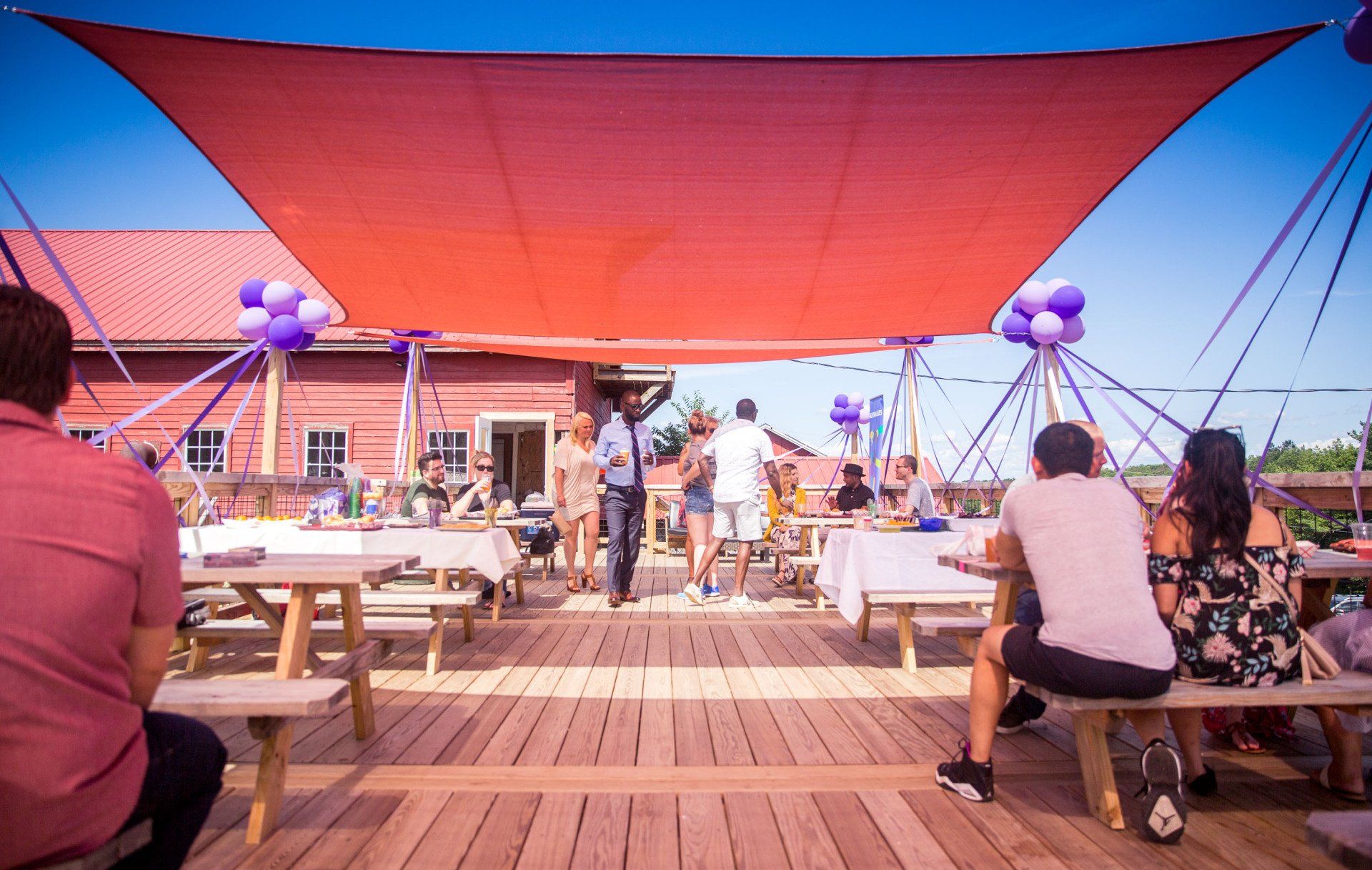 A group of people are sitting at picnic tables under a red umbrella.