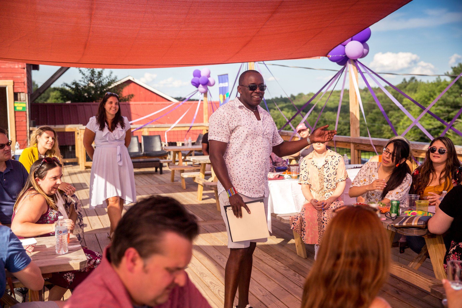 A man is giving a speech to a group of people sitting at tables under a canopy.