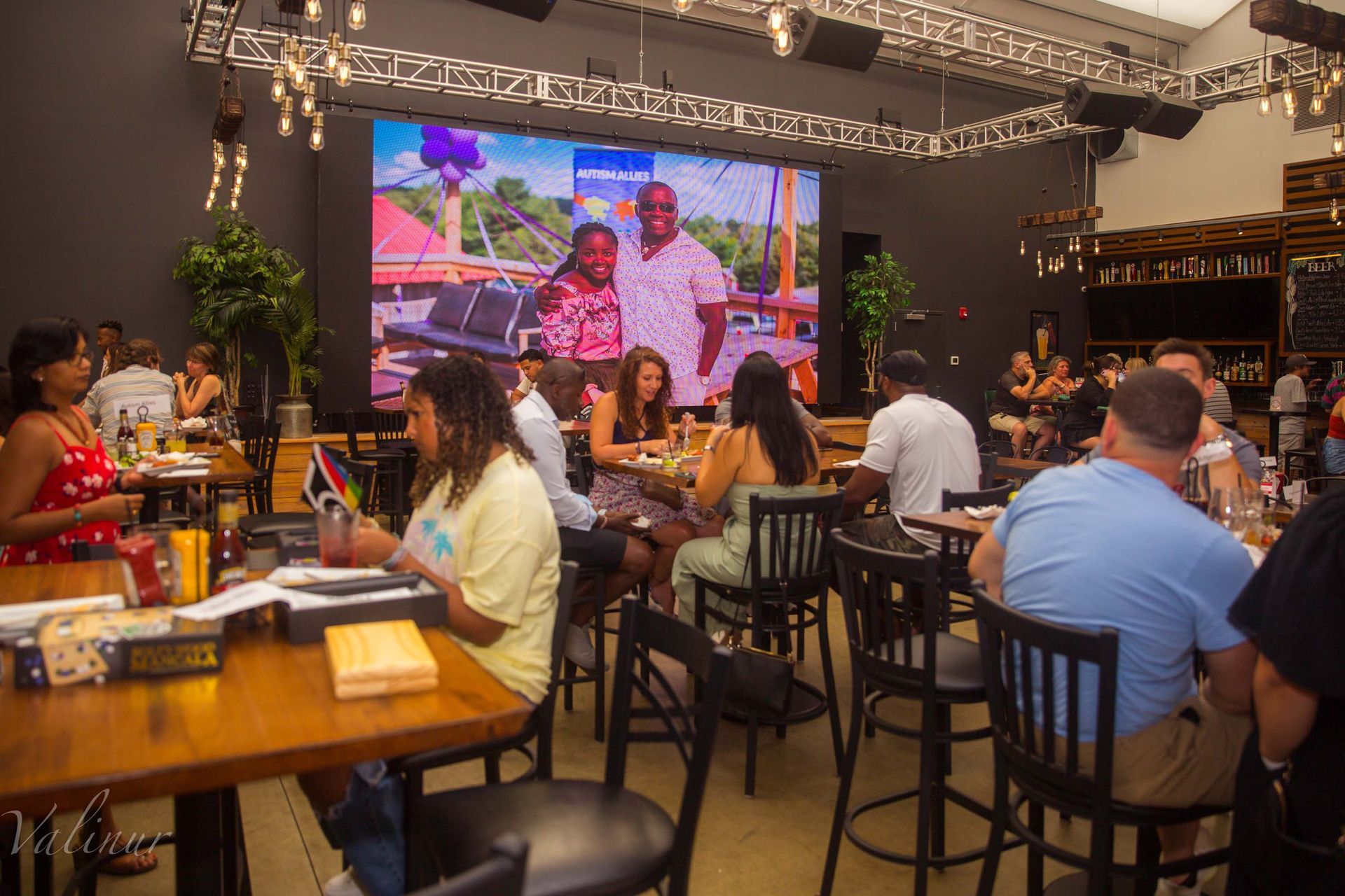 A group of people are sitting at tables in a restaurant in front of a large screen.