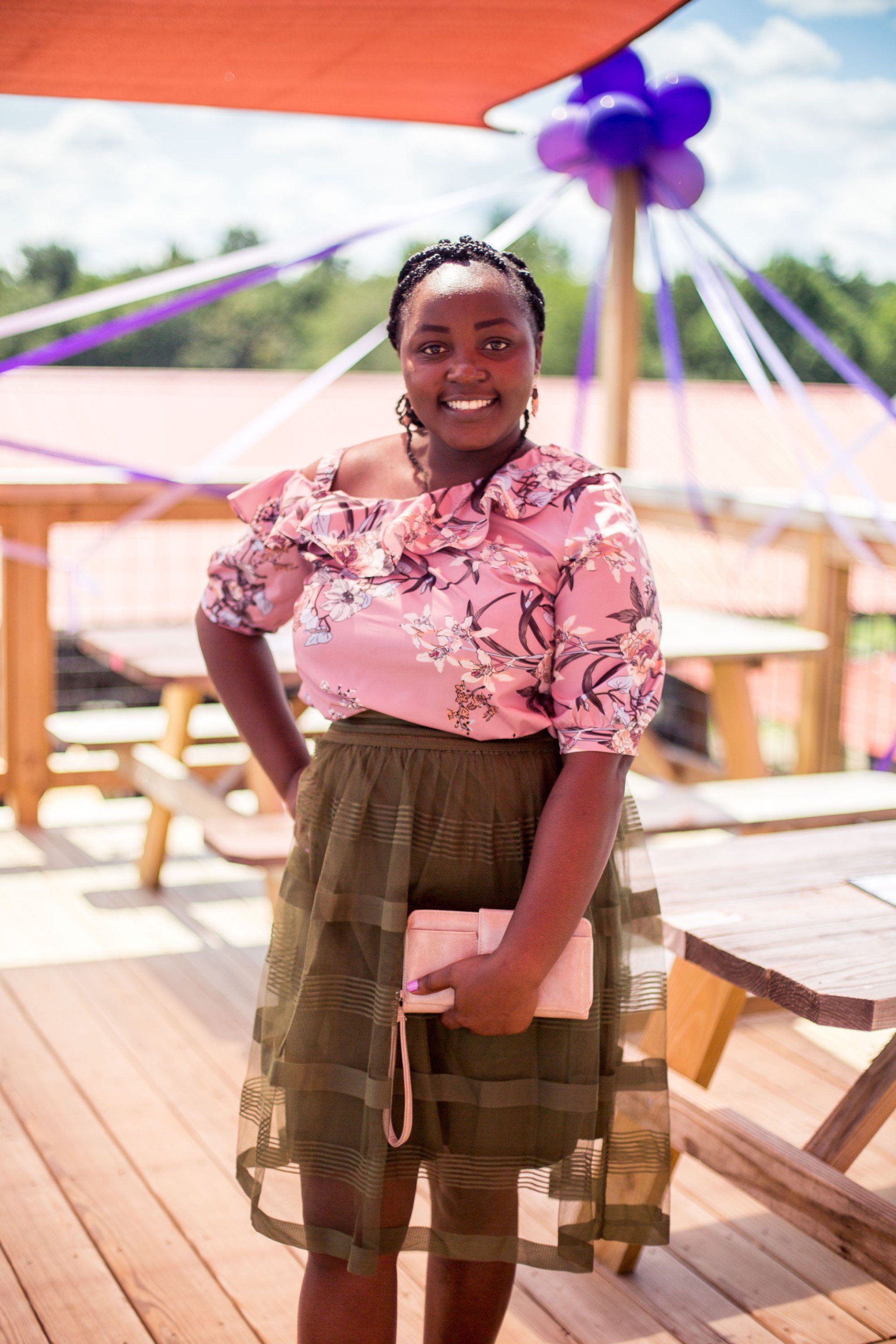 A woman in a pink top and green skirt is standing on a deck holding a pink purse.