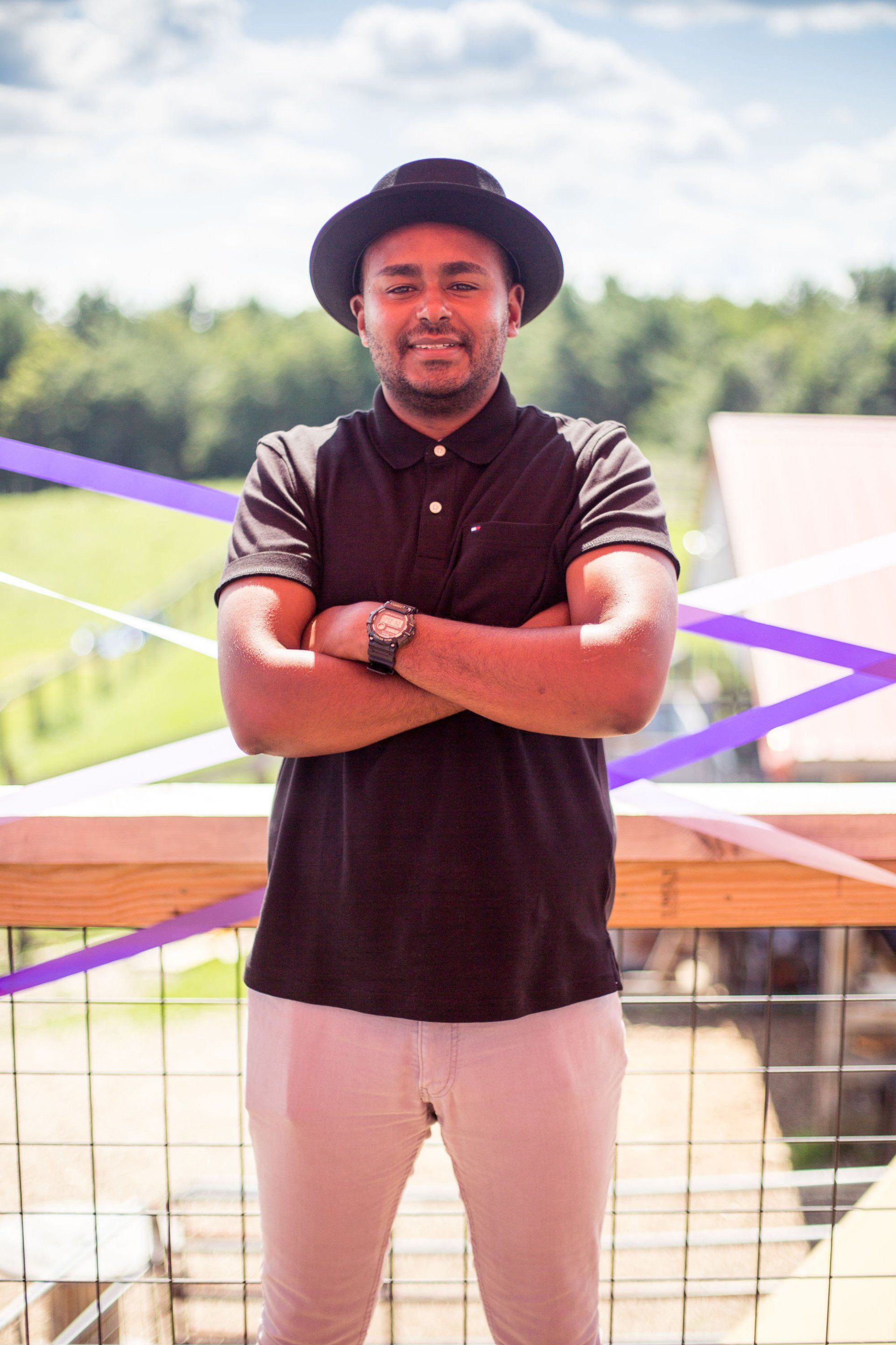 A man wearing a hat and a black shirt is standing with his arms crossed.