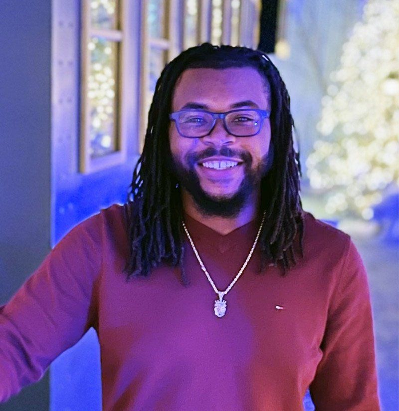 A man with dreadlocks and glasses is wearing a red sweater and a necklace.