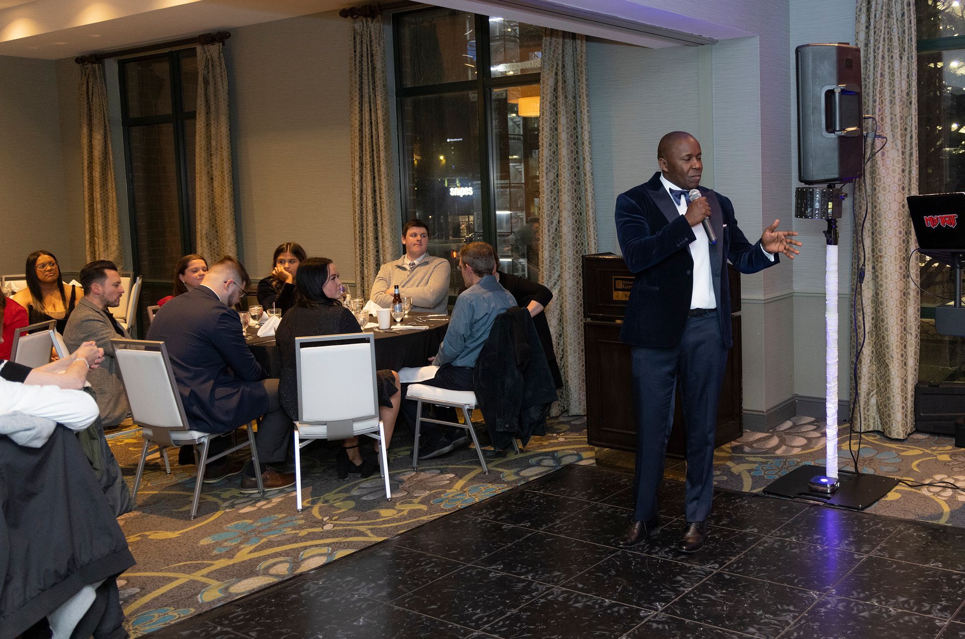 A man is standing in front of a microphone in a room with people sitting at tables.