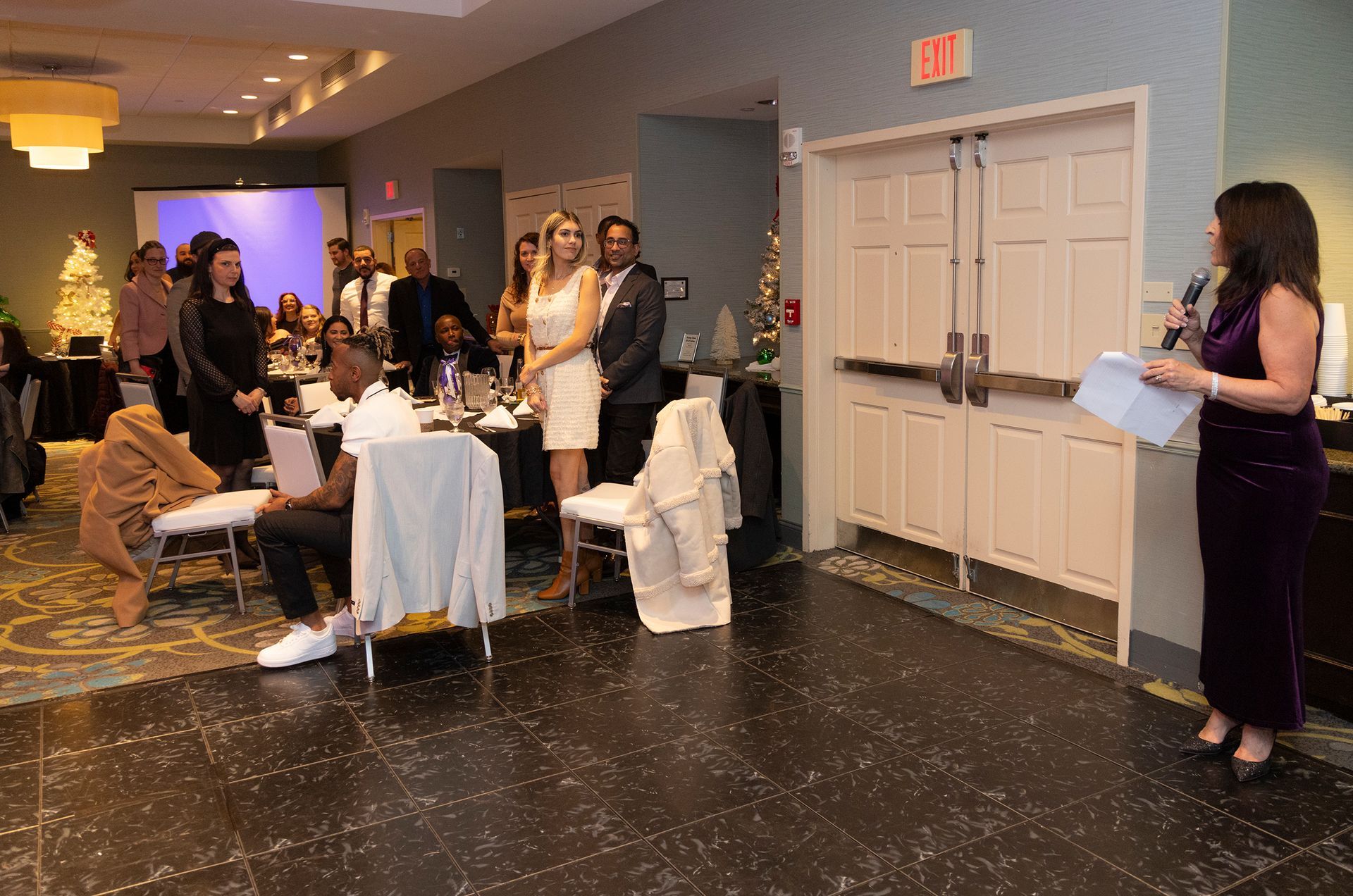 A woman is standing in front of a group of people sitting at tables in a room.