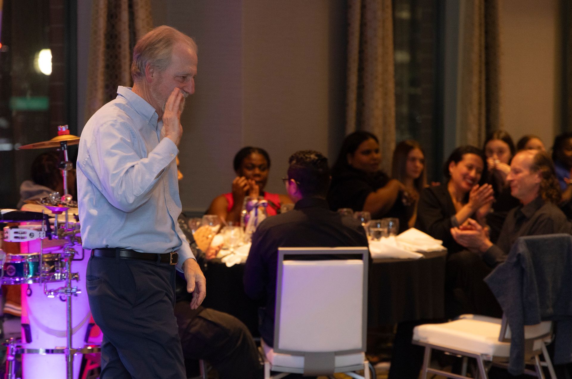 A man is standing in front of a drum set in a room with people sitting at tables.