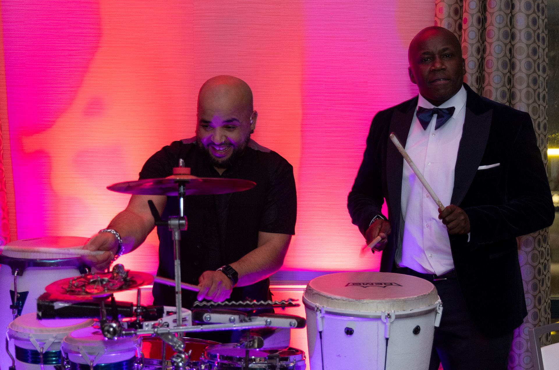 Two men are playing drums in front of a pink wall.