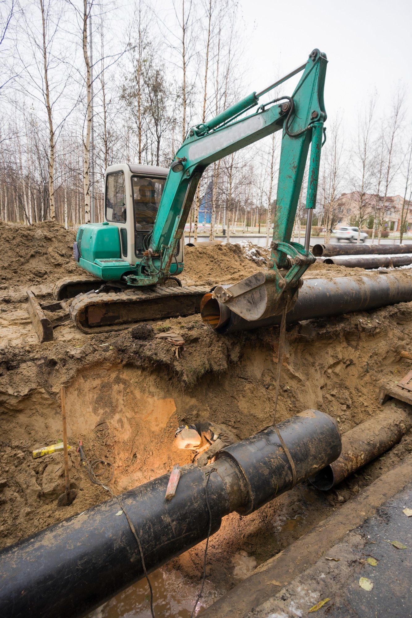 A green excavator is digging a hole in the ground next to a pipe.